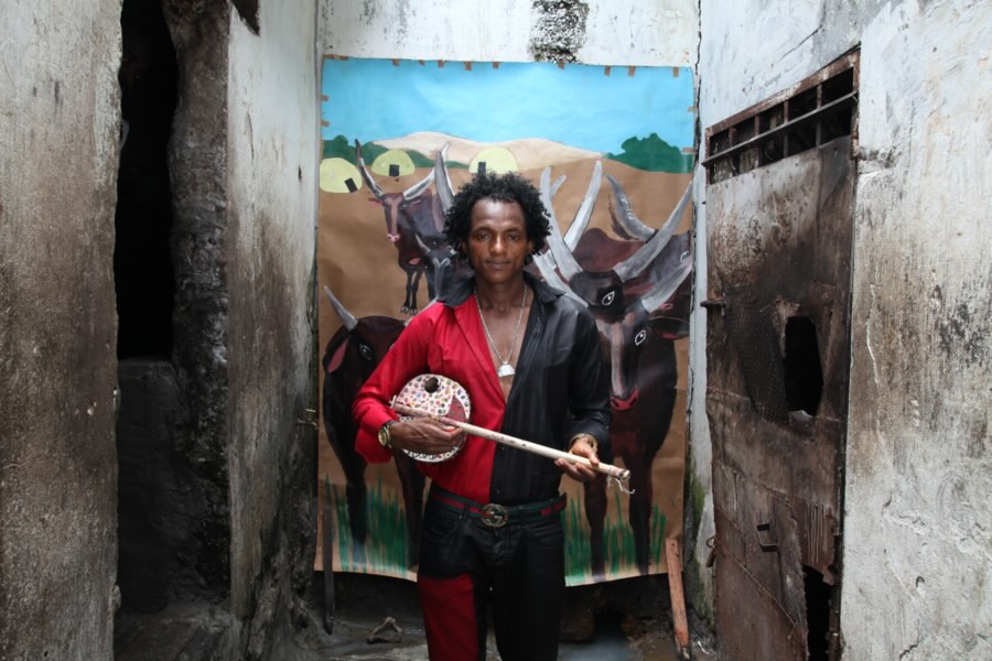 Transporter stands in a narrow courtyard at New Bell Prison holding a long-necked string instrument, in front of a painted backdrop depicting cattle.