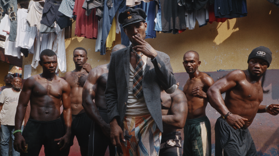 La PJ stands in a blazer, tie, and peaked conductor's cap among a group of men inside New Bell Prison, many of them shirtless, with clothes hanging overhead.
