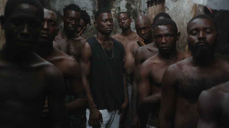 A tightly packed group of men stands in a narrow, weathered passage inside New Bell Prison; most are shirtless, while a man near the center wears a black sleeveless shirt and gold chain.