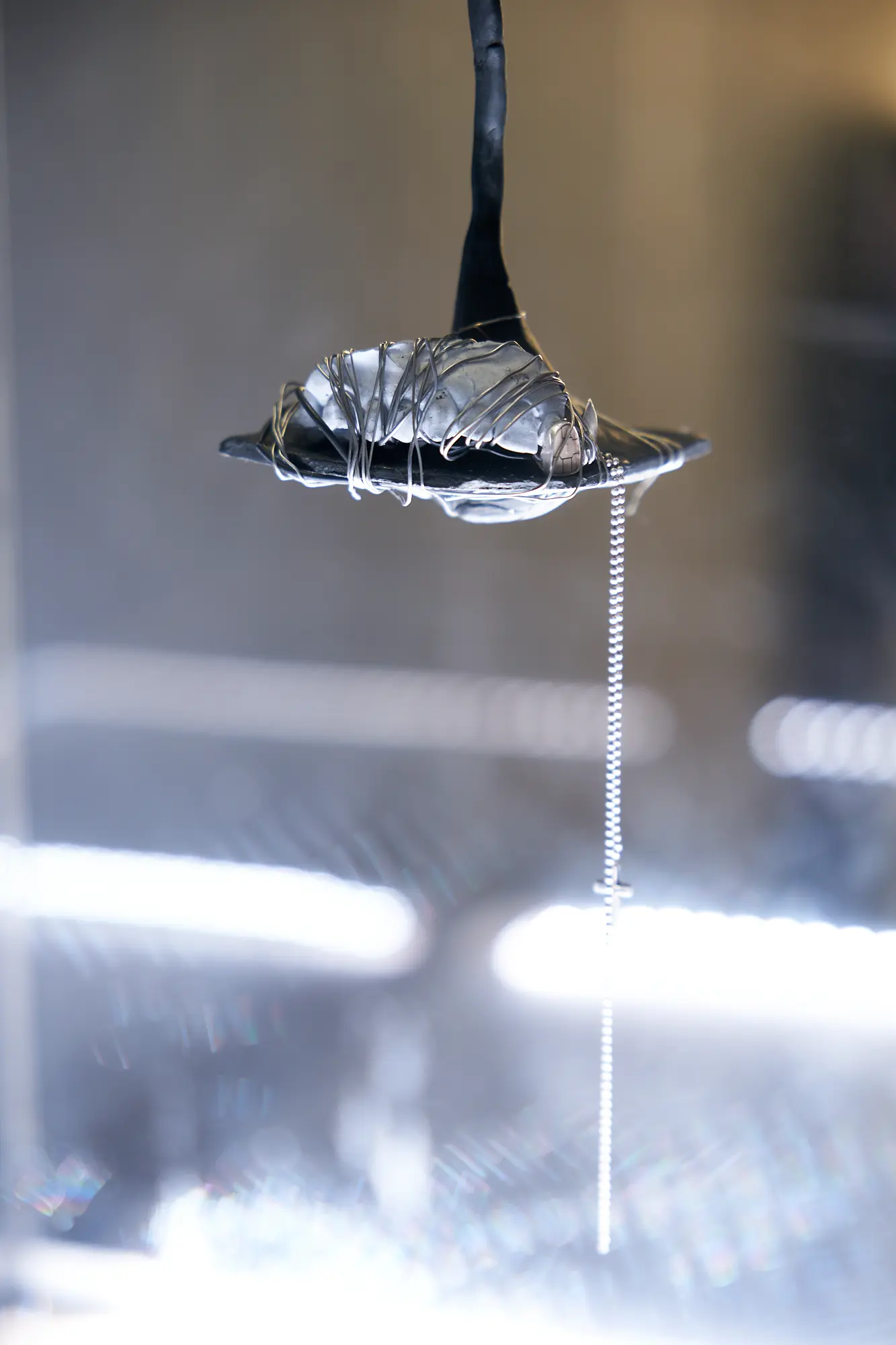 A close-up of a sculptural grill made from a dental mold, wrapped in wire and suspended from a black cord, with a beaded chain hanging below it.