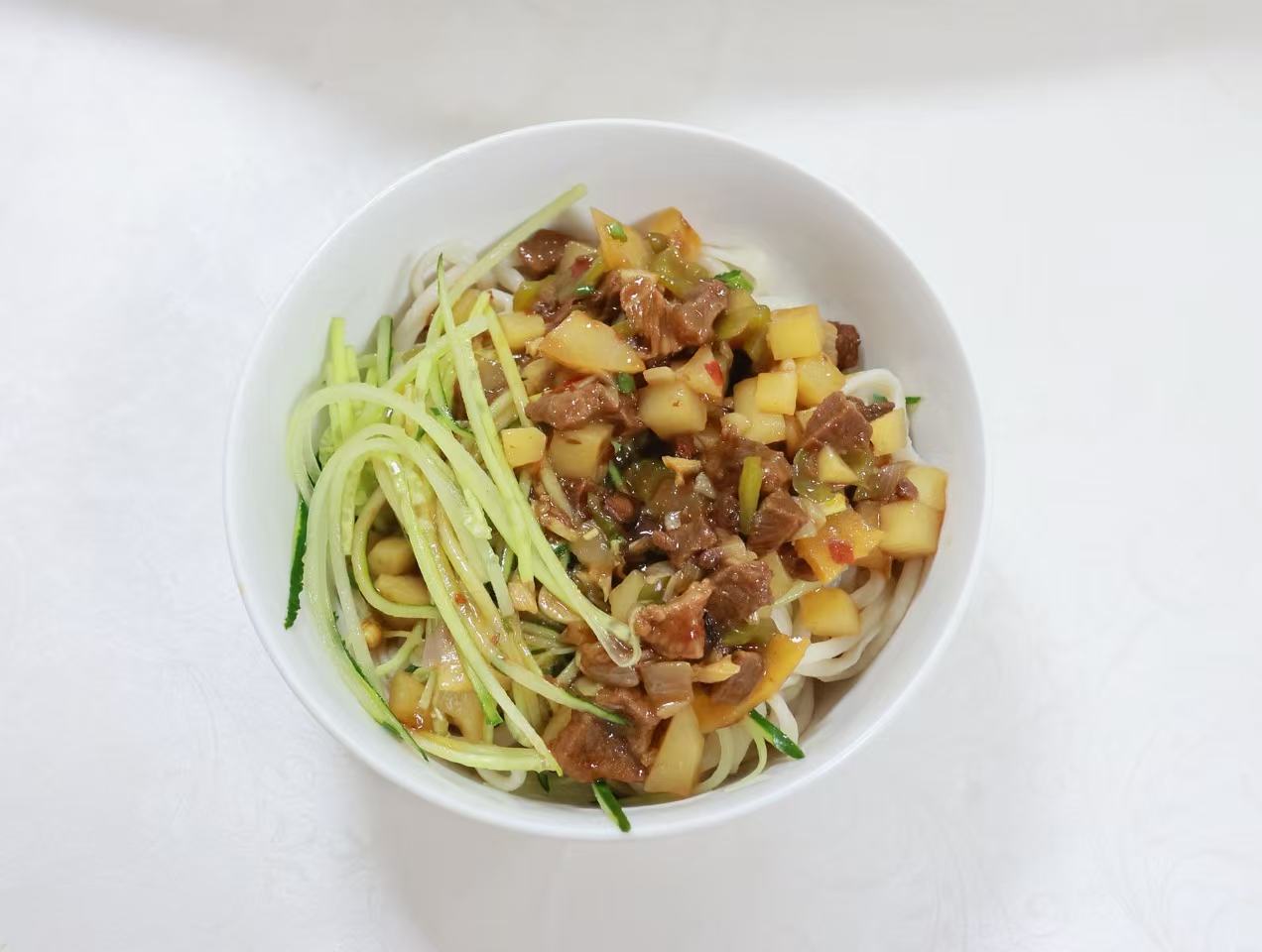 A bowl of hand-pulled Chinese noodles topped with diced beef, vegetables, and julienned cucumber, prepared by a Chadian cook.