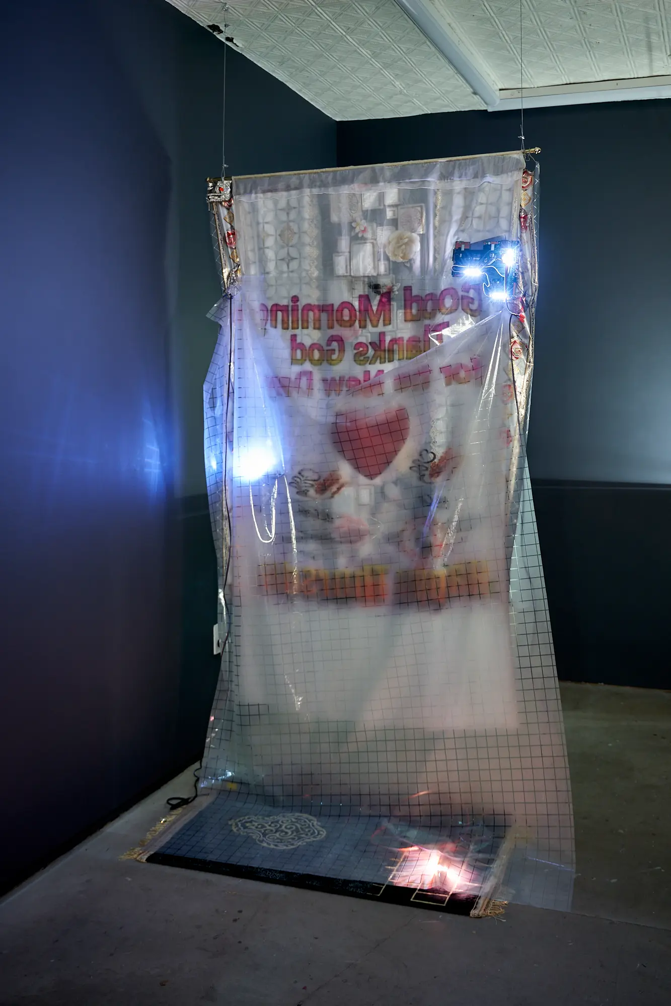 One: an installation view of a translucent banner reading, in mirrored text, “Good Morning Thanks God for the New Day,” hung from a gallery ceiling and lit by small flash units, above a prayer rug.