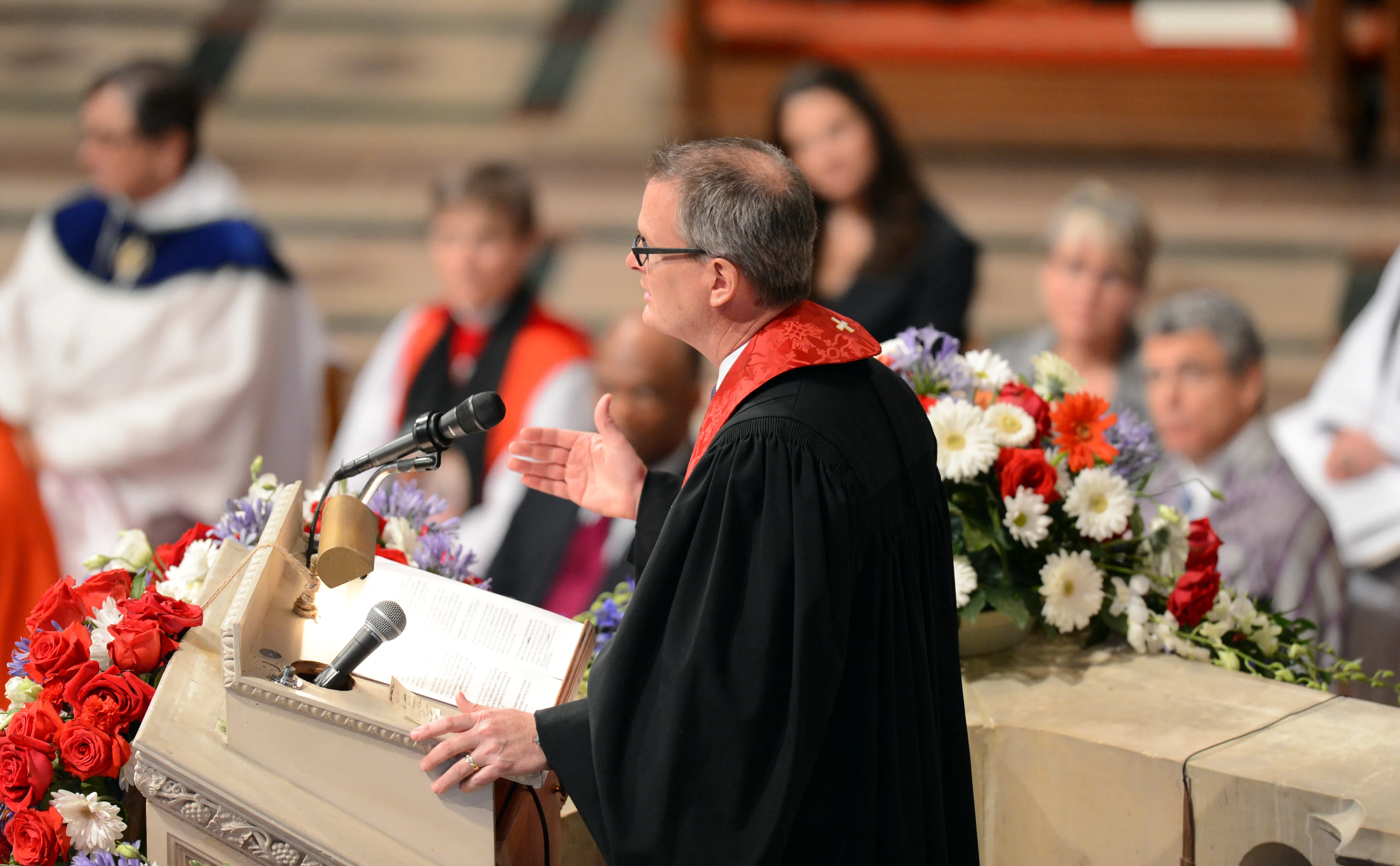 A man in a black robe and red stole addresses an audience from a pulpit adorned with red and white flowers. An open book rests in front of him.